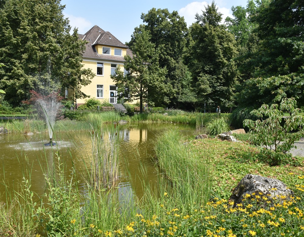 Ein weißes, zweistöckiges, älteres Haus mit rotem Dach steht inmitten einer grünen Parklandschaft und vor einem Springbrunnen mit Fontäne