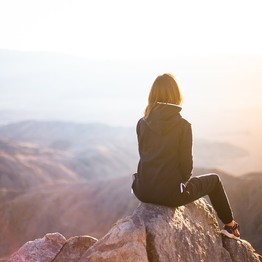 Eine Frau in dunkler Kleidung sitzt auf einem Berggipfel und schaut in die Landschaft