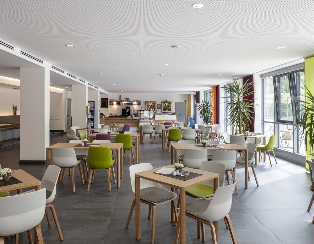 Blick in den modernen Innenraum des Cafés, mehrere Tischgruppen mit weißen und grünen Stühlen, grünen Pflanzen am rechten Bildrand zur Fensterfront